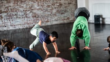 A child and teacher are both in downward facing dog (yoga pose). The child is kicking one of their legs up and looking at the rest of the class.