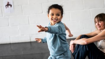 A smiling child reaches forwards as they follow along in a dance class.