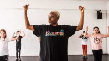 A teacher stands in front of a class of children with their arms raised to shoulder height and bent at right angles. The children are copying the same pose.