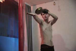 A topless performer pours a liquid from a pan onto themselves, in soft pink lighting