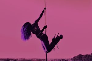 A dancer with long curly hair and stiletto heels hangs from a pole against a pink sly