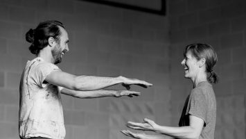 two laughing dancers stand facing each other, palms outstretched