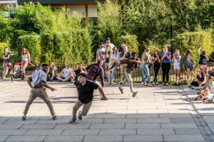Four dancers from different ethnic backgrounds running and smiling in the centre of a large square. The audience are spread along the edges watching and smiling.