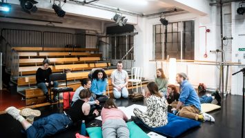 A group of people of many ages sit in a circle on the floor of a dance studio, deep in discussion