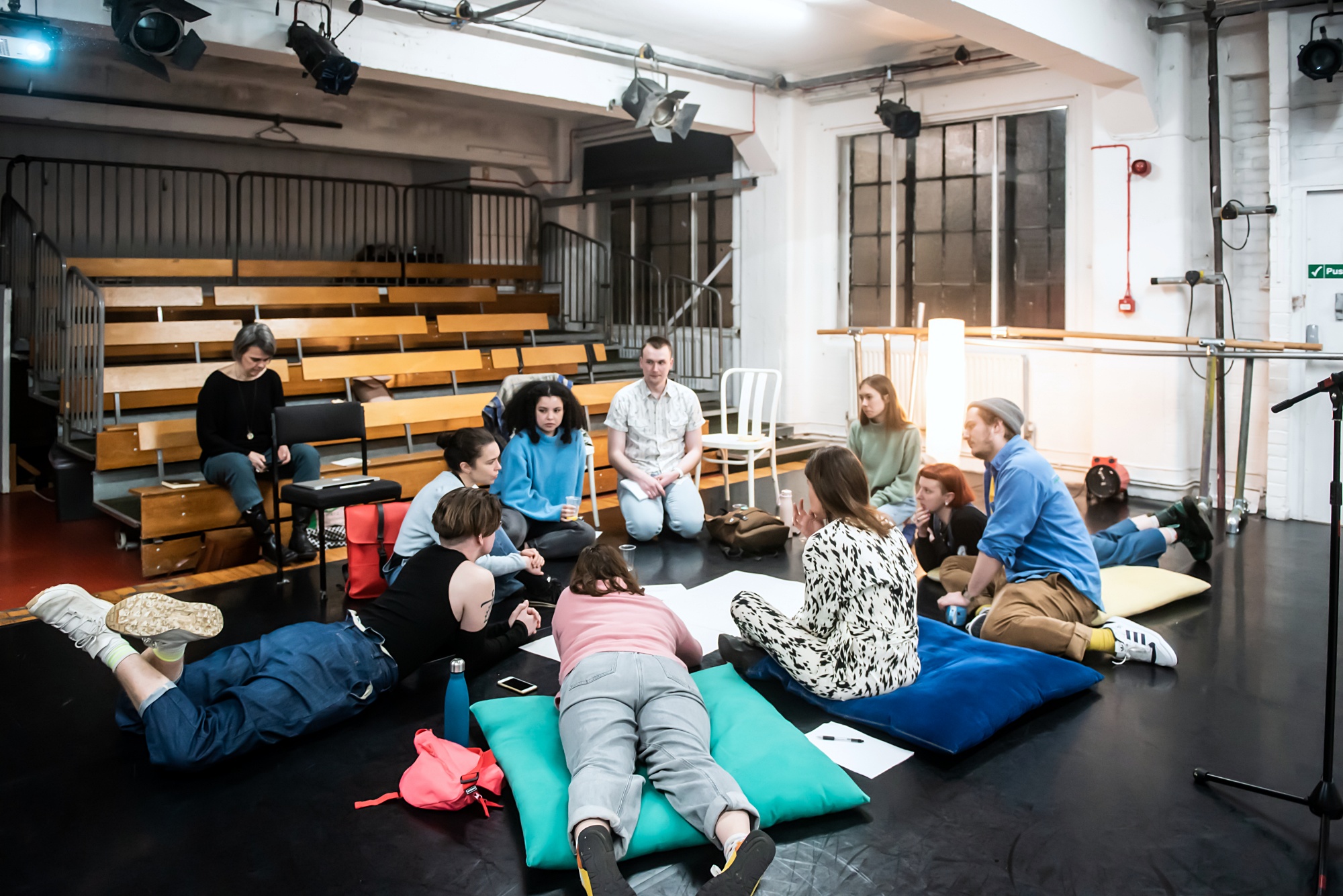 A group of people of many ages sit in a circle on the floor of a dance studio, deep in discussion