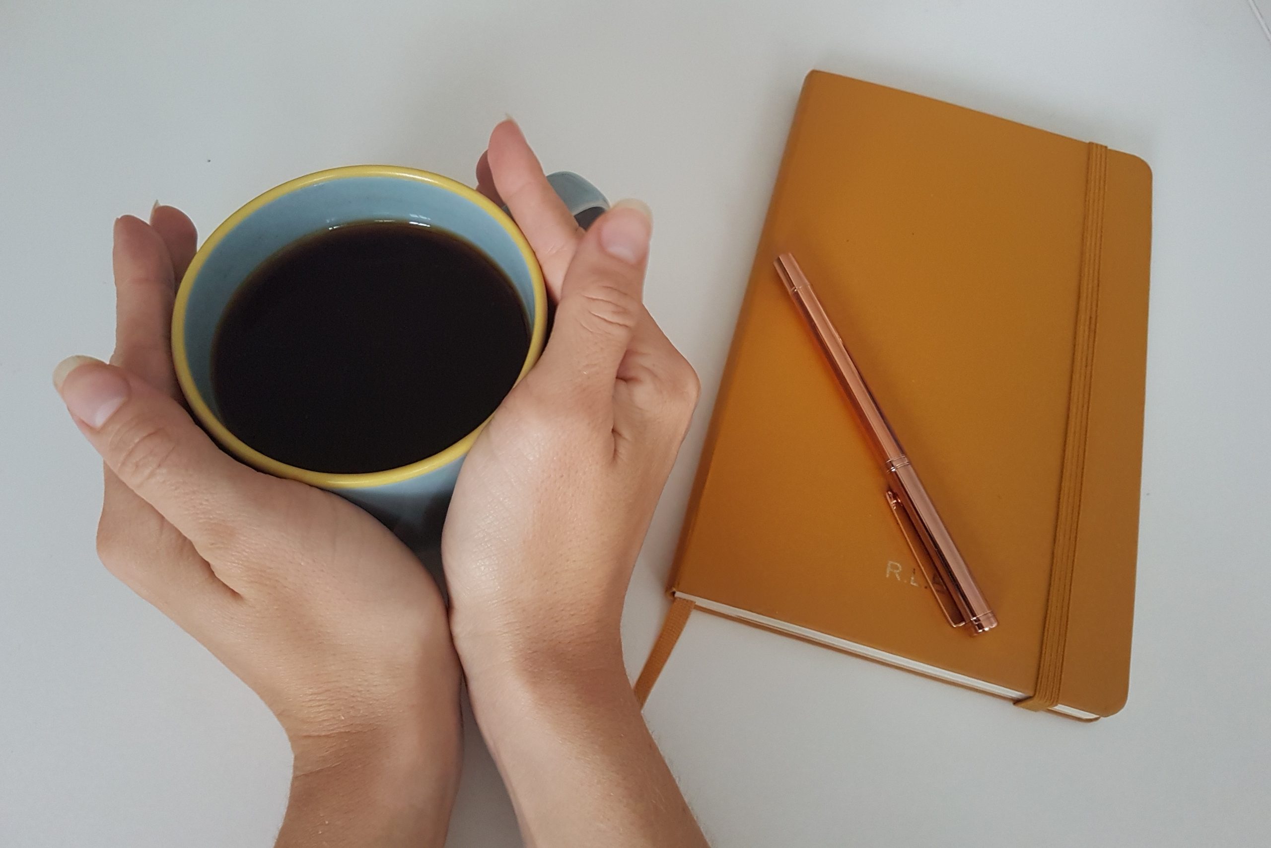 A pair of hands wrap around a mug of coffee, next to a notebook and pen
