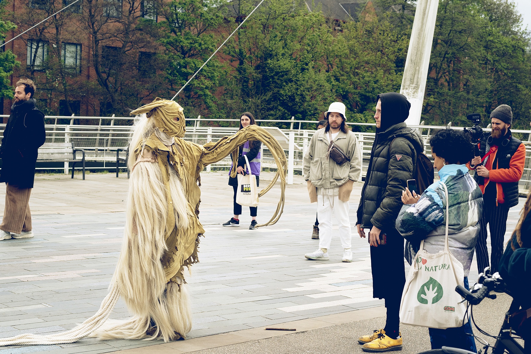 a masked performer approaches an audience member outdoors in daylight. They outstretch a long curled arm and are wearing a beige/white costume covered in long white hair and beige fabric