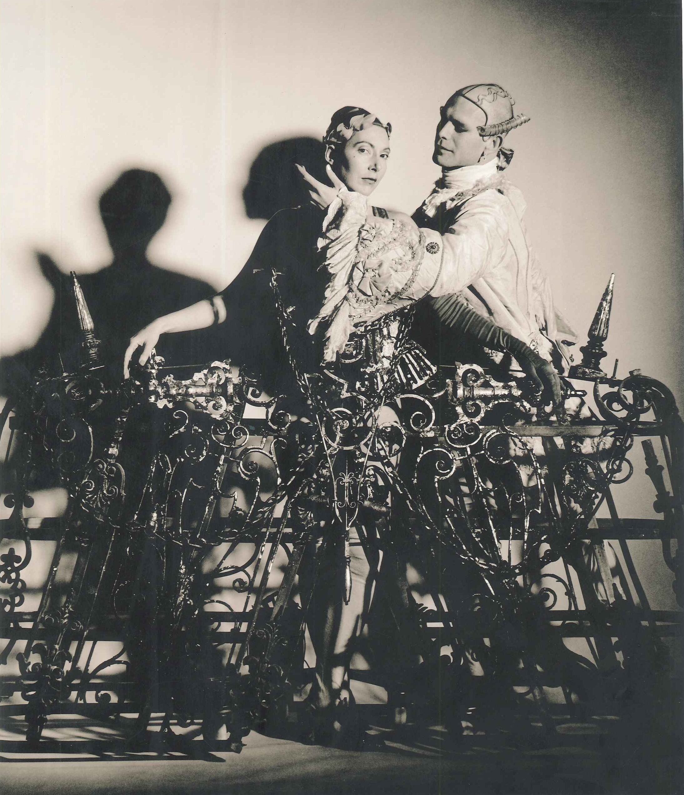 A sepia image of two performers dressed in ornate ruffled costumes stand delicately inside an elaborate waist-height cage