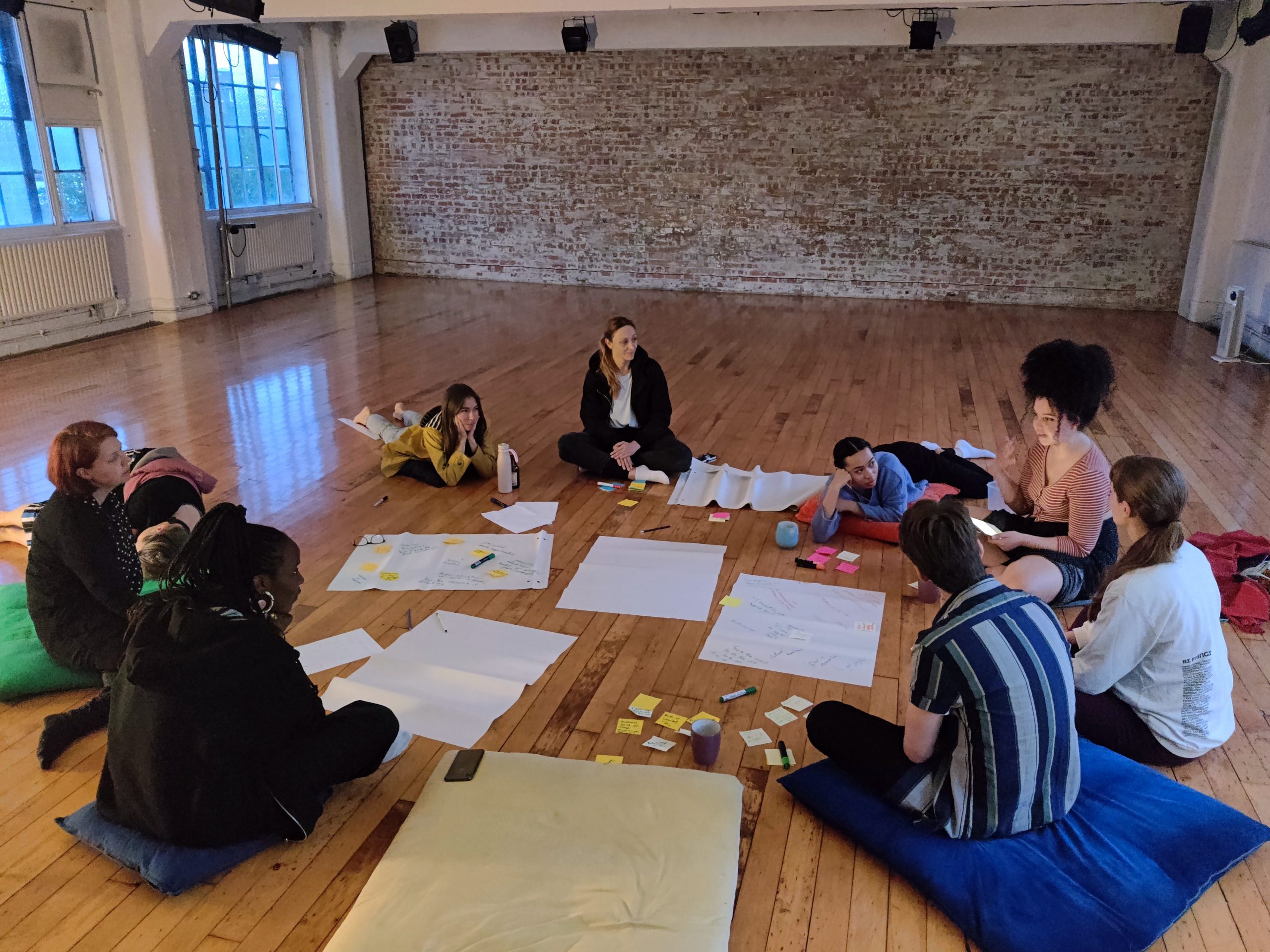 A group of artists sit in a circle on the wooden floor of a dance studio in the early evening. A mixed race woman is leading a conversation whilst they sit on large cushions. There are big sheets of paper on the floor between them