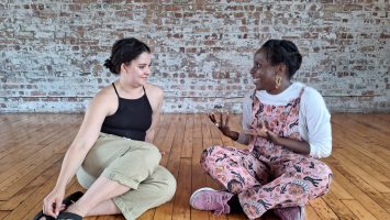 Two artists are having a conversation, sat on a wooden dance floor. There is a brick wall behind them.