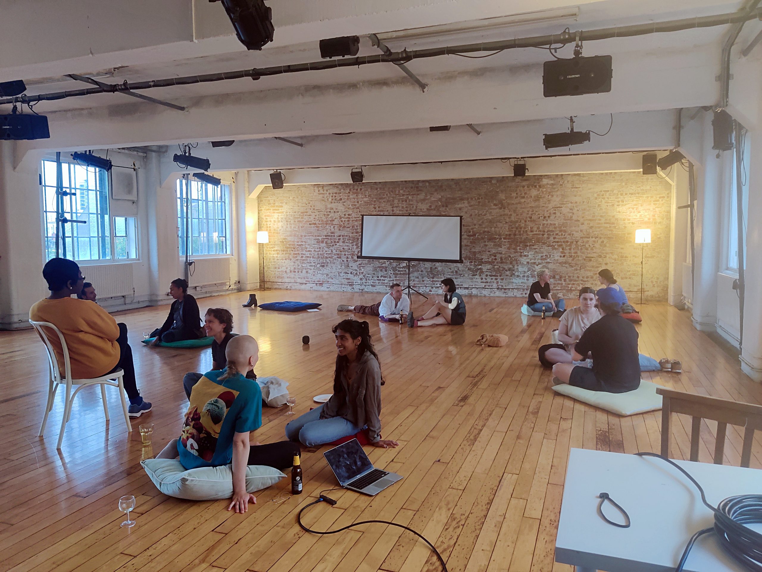 Around 15 artists sit on floor cushions and in chairs, in a warmly lit studio. They are in pairs deep in discussion, several are smiling.