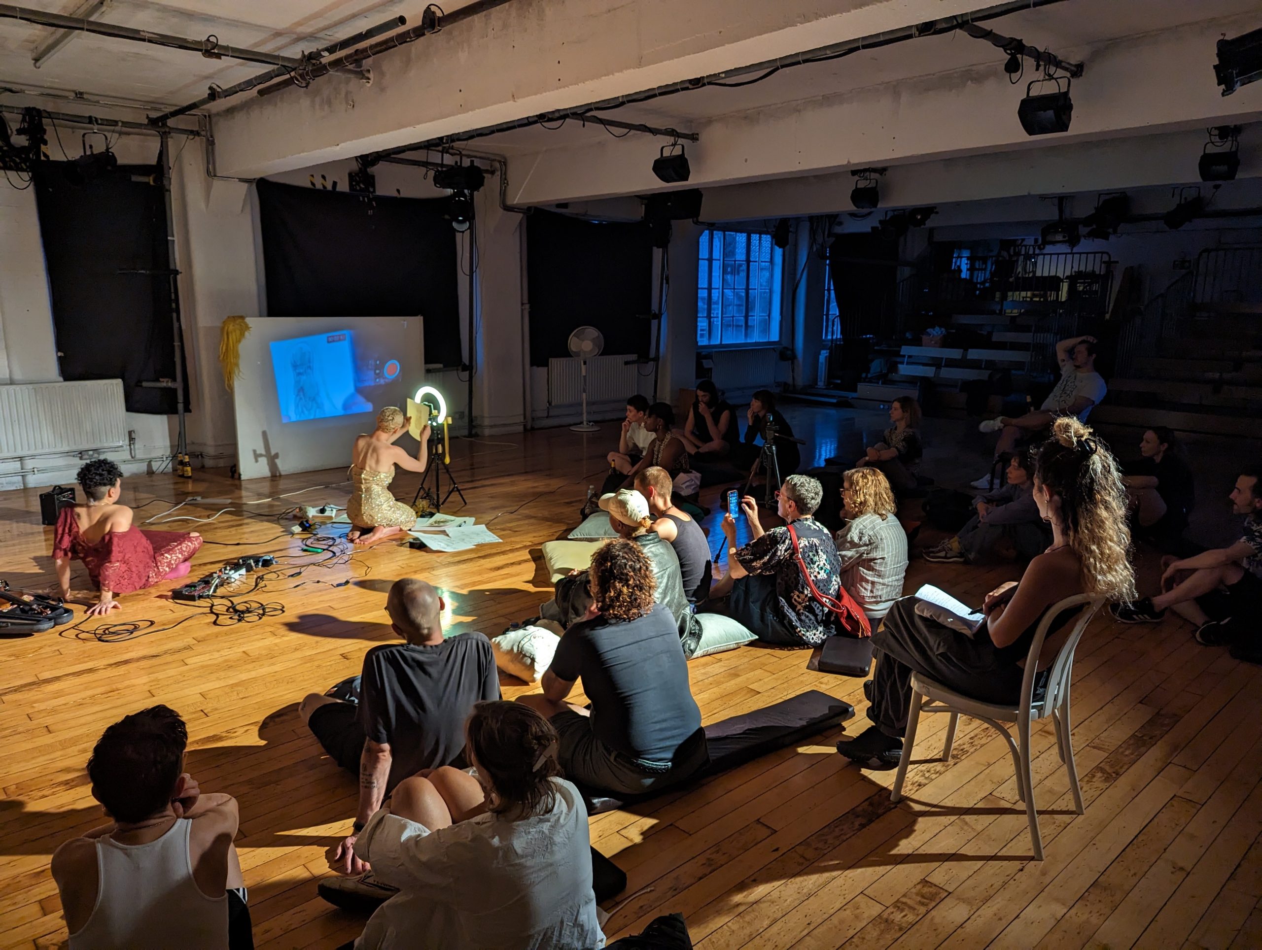 A photo of a performance event, dimly lit in a studo with a wooden floor at dusk. Two light skinned artists with short hair sit amongst a complicated array of technology - cables, sound equipment, a phone on a tripod, and a live video feed to a projector screen.